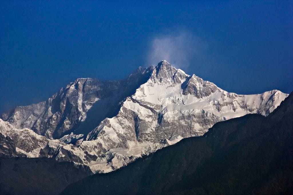 A panoramic view of Mount Kanchenjunga in India, with its snow-capped peaks and a small puff of cloud or smoke beneath a blue sky.