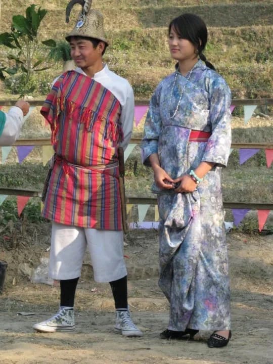 A young Lepcha man and woman in traditional Bhutanese or Himalayan costume standing in an outdoor setting, the man wearing a striped gown and hat, while the woman wears a blue kimono-style dress and necklace.